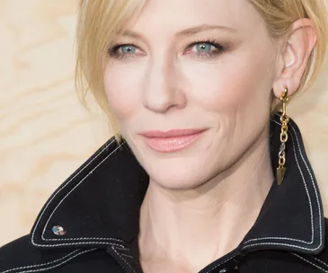 A close-up of a person with blonde hair, wearing a dark jacket and gold earring, looking slightly to the right.