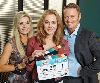 "Three actors from 'The Wrong Girl' Season 2 smiling, one holding a clapperboard, on set during filming."