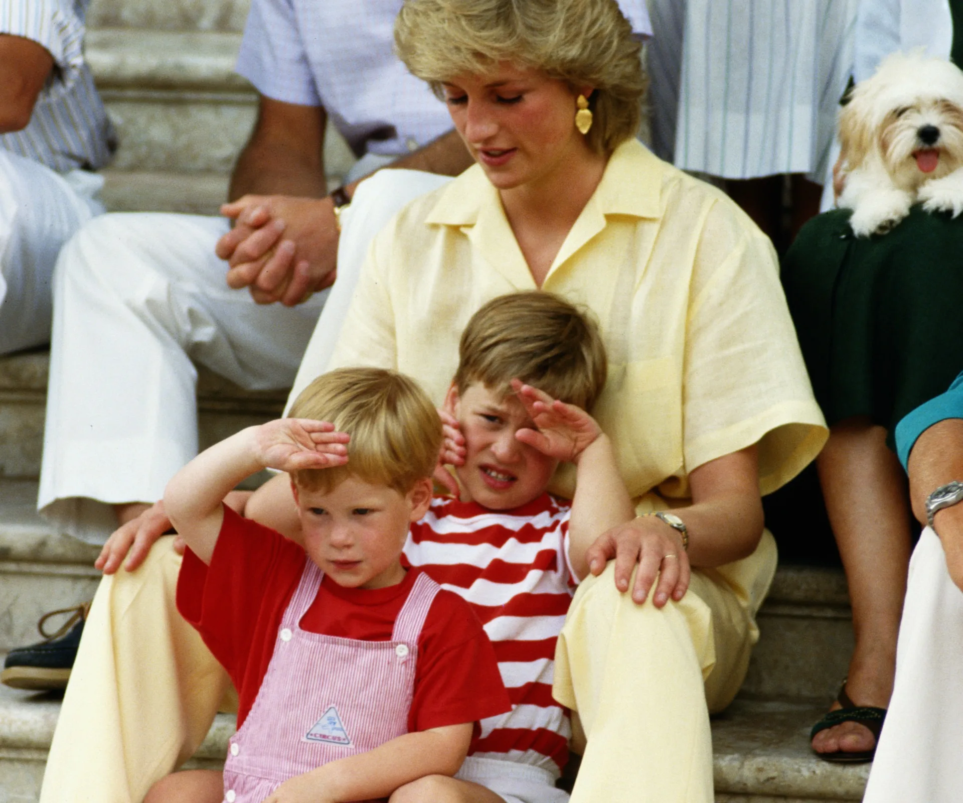 A woman sitting on steps with two young children, one in red overalls, a dog is beside them.