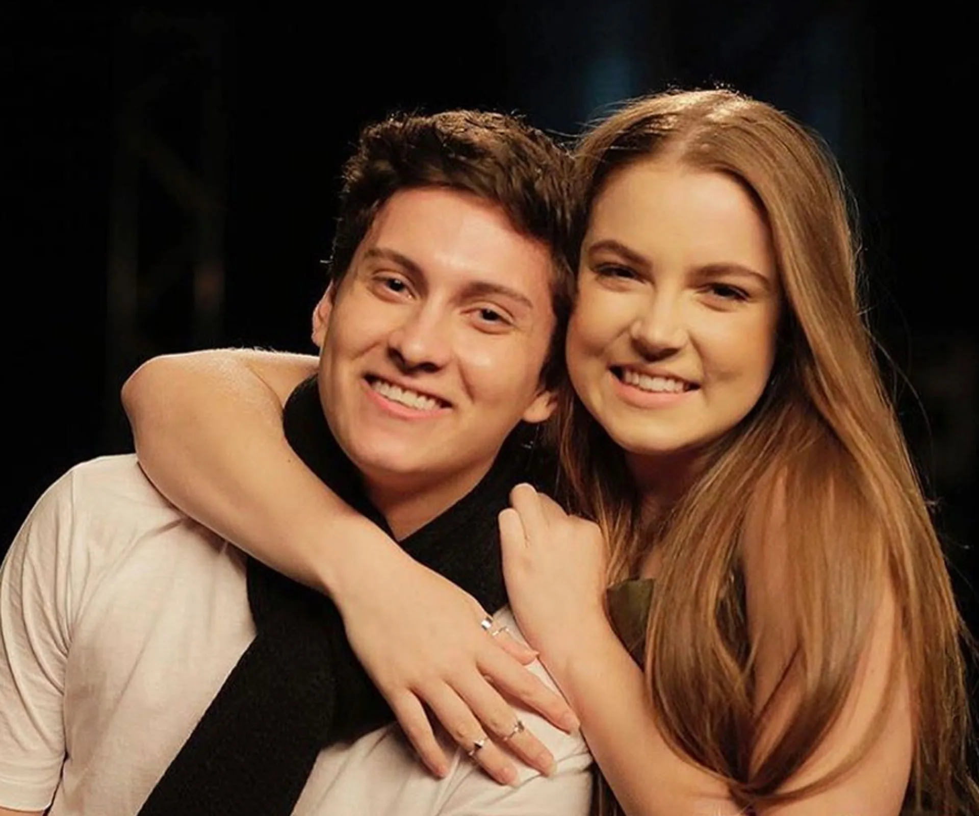 A smiling young woman hugs a smiling young man from behind, both looking at the camera.