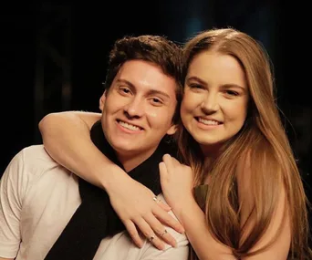 A smiling young woman hugs a smiling young man from behind, both looking at the camera.