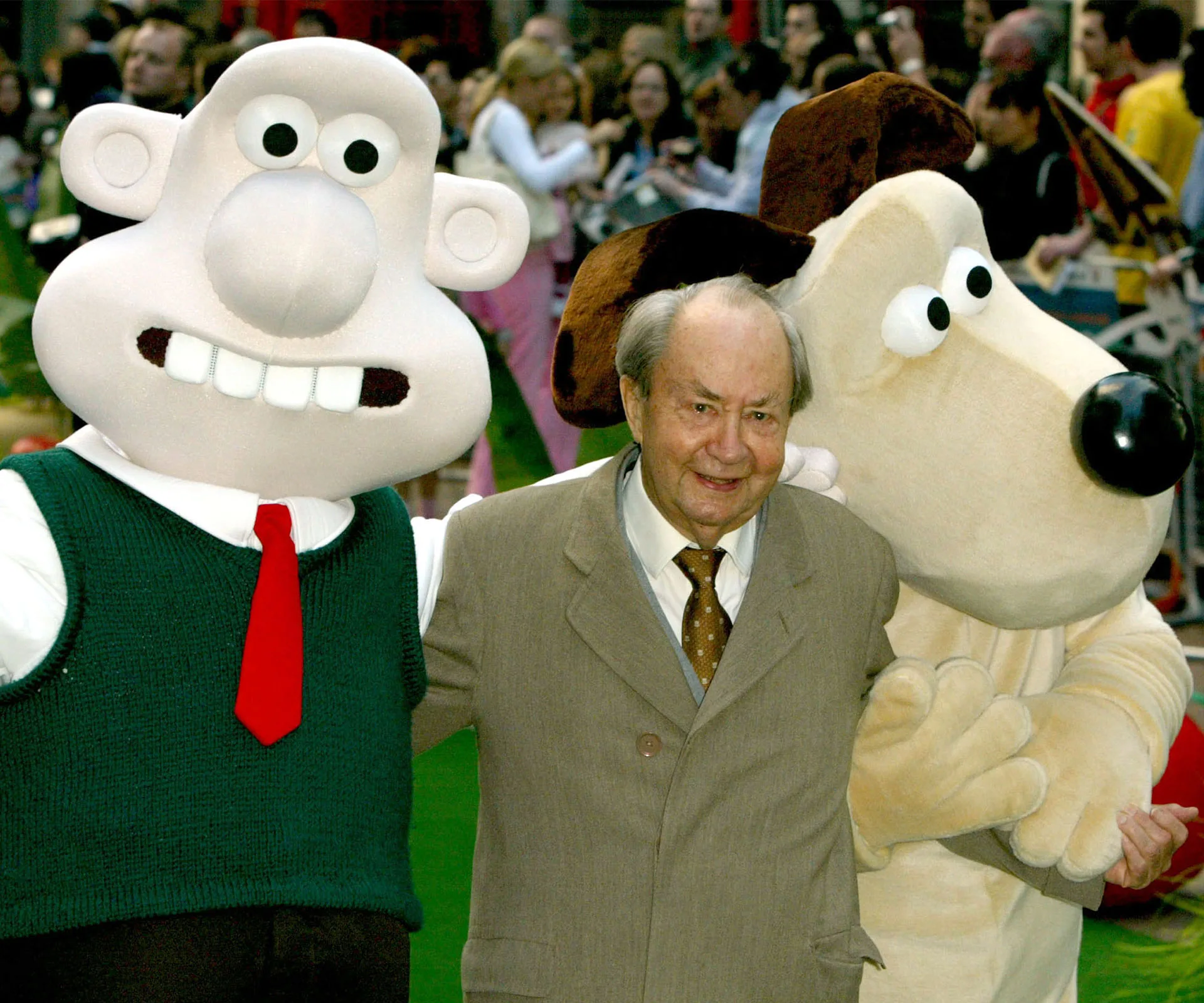 Peter Sallis with Wallace and Gromit mascots at an event, smiling and posing for a photo.