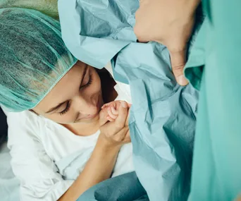 Mother wearing surgical cap holding newborn's hand during C-section, smiling lovingly.