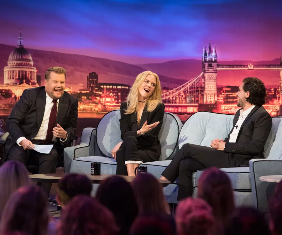 James Corden, Nicole Kidman, and Kit Harington laugh on a talk show set with a London backdrop.