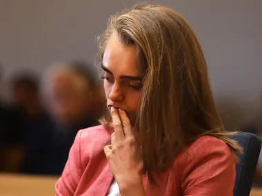 A person in court, appearing pensive, with hand on face, wearing a pink blazer.