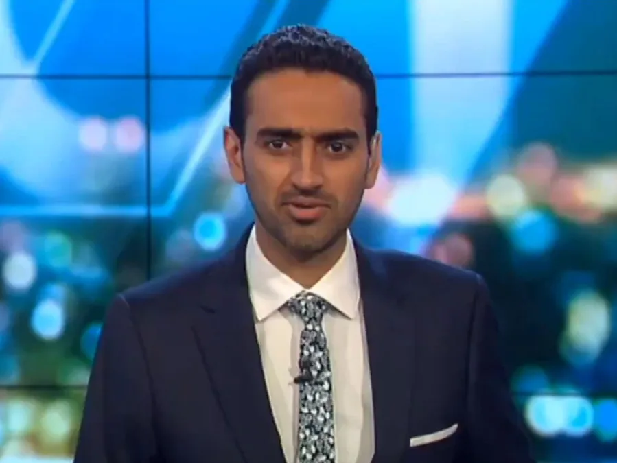 A man in a suit and patterned tie stands in front of a colorful, blurred background on a television set.