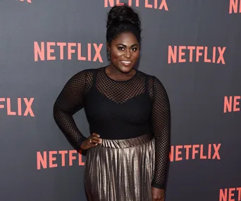 Danielle Brooks posing at a Netflix event, wearing a black mesh top and metallic skirt, smiling against the Netflix backdrop.