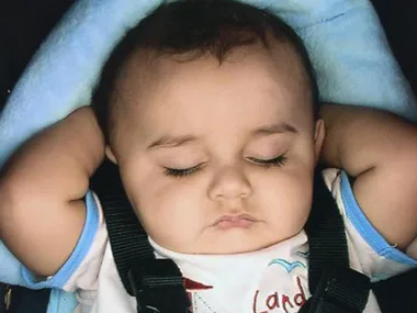 A sleeping baby in a car seat with arms behind head, wearing a white shirt with blue edging.