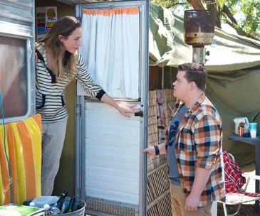 A woman leans out of a caravan door, talking to a young man in a plaid shirt outdoors.