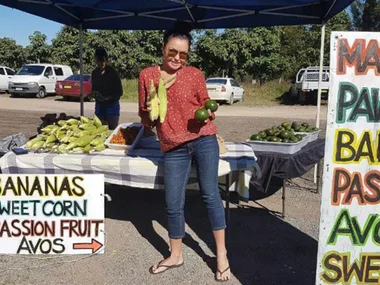 Person at a market stall holds sweet corn and avocados, with signs displaying available produce.