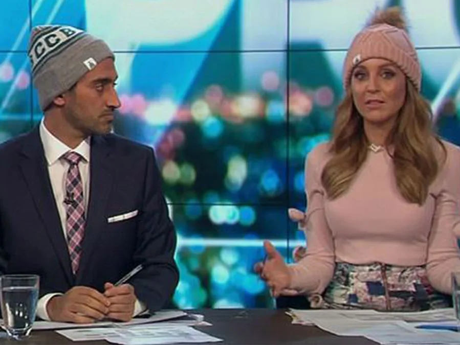 Two television presenters are wearing beanies while sitting at a desk on a studio set.