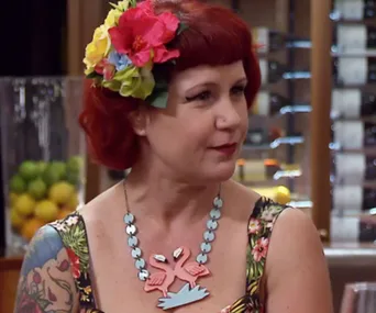 Red-haired woman with flower hair accessory, floral dress, bird necklace, and arm tattoo in restaurant setting.