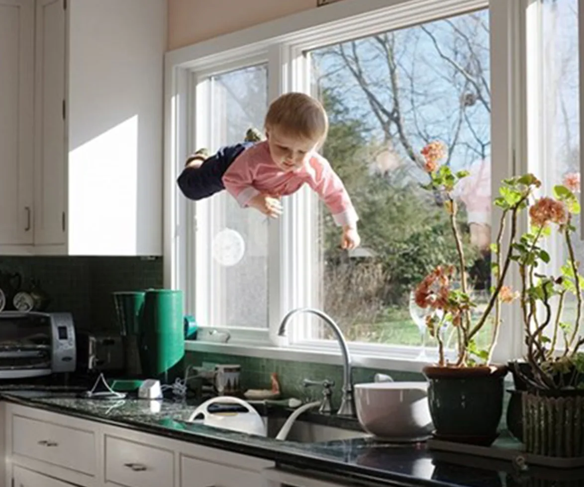 A baby wearing a pink sweater flies horizontally indoors, with a kitchen sink and potted plants beneath.