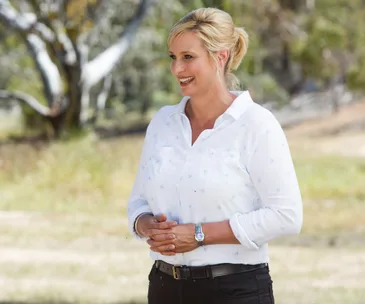 Host in a white shirt smiles outdoors with trees in the background.