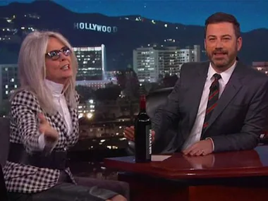 "Person wearing glasses and checkered blazer talks animatedly on a late-night show set with another person in a suit."