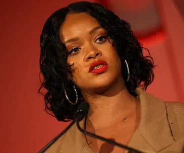 Singer speaking at an event with a serious expression, wearing red lipstick and hoop earrings.
