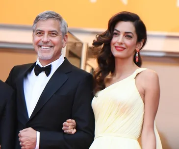 A couple in formal attire smiles on a red carpet event, the man in a tuxedo and the woman in a yellow dress.