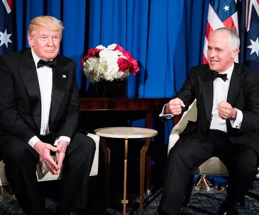Two men in tuxedos sit in chairs, with blue curtains and flags in the background, one gesturing while the other listens.