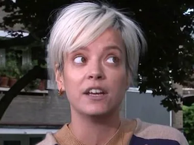 A person with short blond hair talking outside, with trees and buildings in the background.