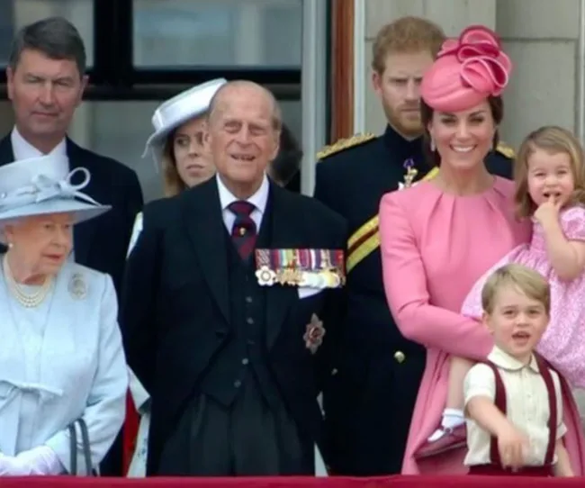 People stand on a balcony during Trooping the Colour, with some in formal attire and military uniforms.
