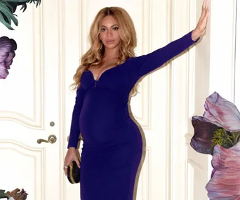 Pregnant woman in a form-fitting purple dress poses by a floral-decorated wall.