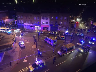 Aerial view of police vehicles with flashing lights at a night crime scene in London, with a crowd gathered nearby.