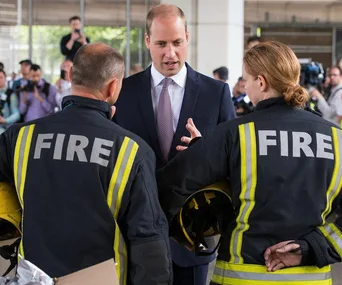 A man in a suit converses with two firefighters wearing uniforms labeled "FIRE," amid a crowd and photographers.