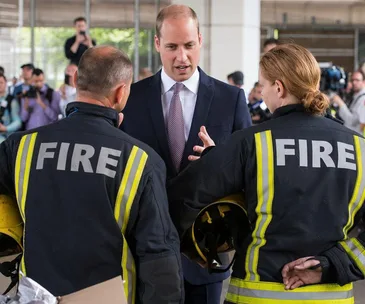 A man in a suit converses with two firefighters wearing uniforms labeled "FIRE," amid a crowd and photographers.