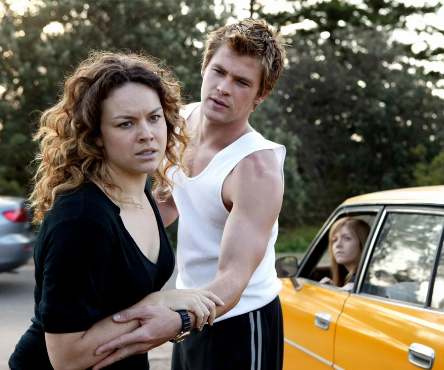 A man holds a concerned woman by the arm; another woman watches from a yellow car in the background.