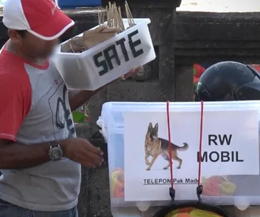 A vendor in Bali holds a container labeled "Sate" with skewers, next to a box with a dog image and "RW Mobil" sign.