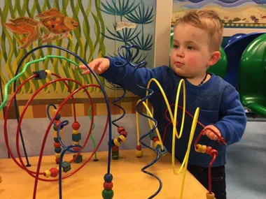 Child playing with a bead maze toy at a colorful play area with a fish mural on the wall.