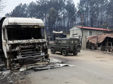 Burned truck and damaged buildings in forest fire aftermath in Portugal, with a vehicle driving past.