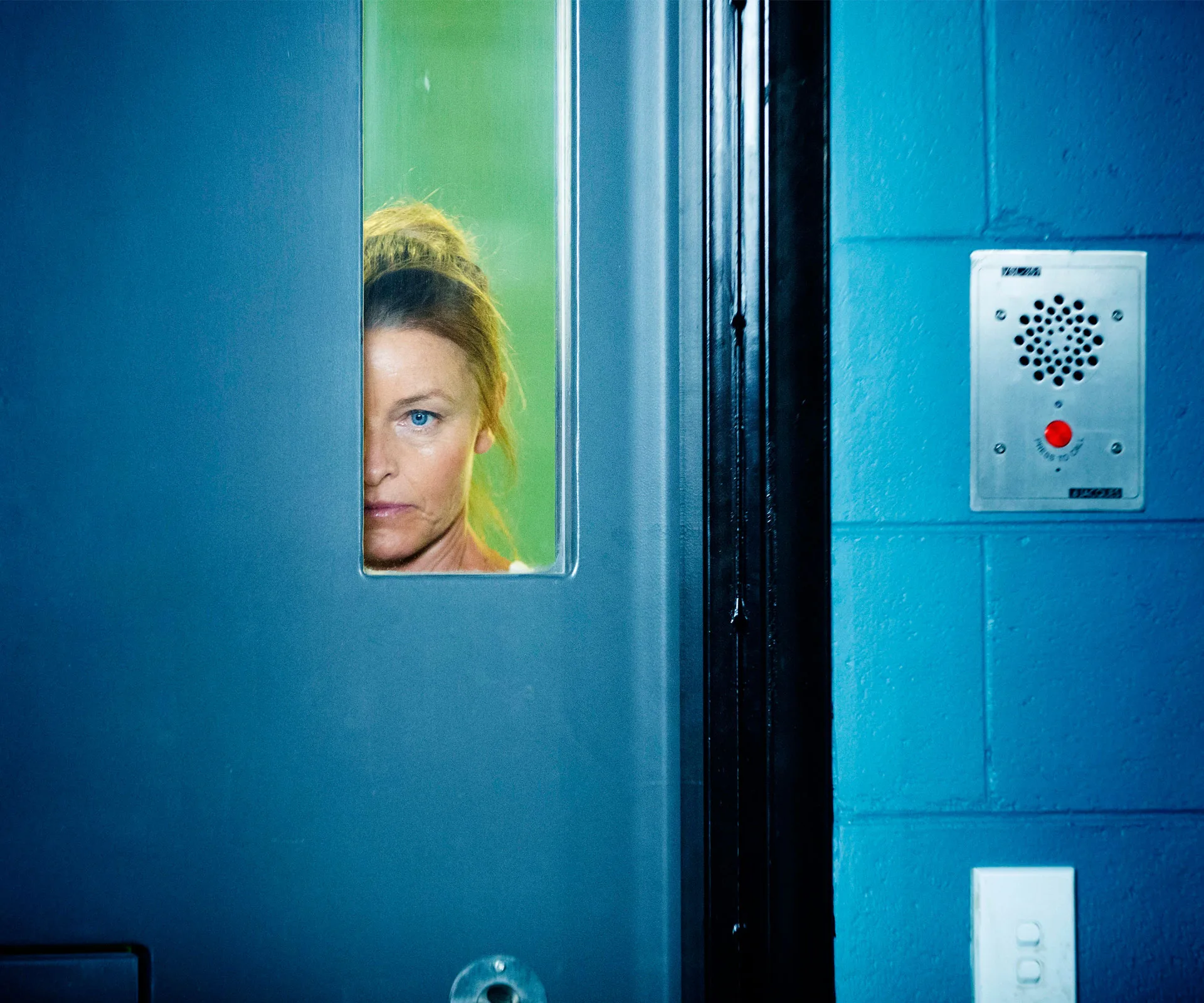 A woman peers through a narrow window in a blue prison door, with an intercom panel nearby.