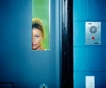 A woman peers through a narrow window in a blue prison door, with an intercom panel nearby.