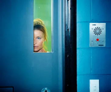 A woman peers through a narrow window in a blue prison door, with an intercom panel nearby.