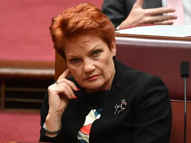 A woman with short red hair and a black outfit sits in a legislative setting, looking attentive.