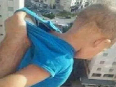 A baby in a blue shirt is held by the back of the shirt out of an apartment window with buildings below.