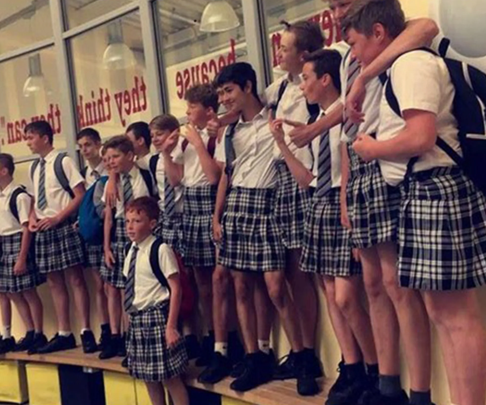 Schoolboys in ties and plaid skirts protest heatwave, standing together indoors.