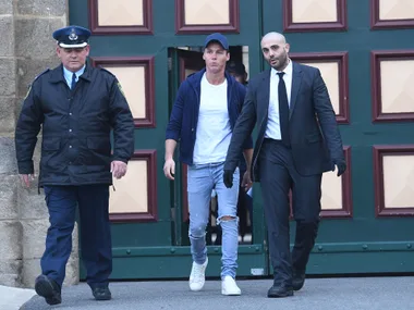 Man in cap with security personnel, walking away from large green door.