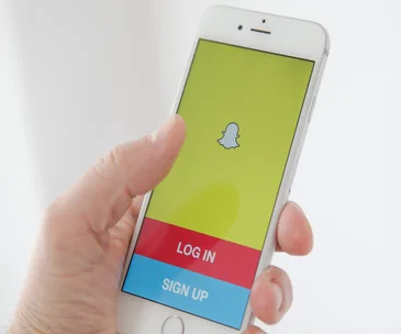 Hand holding a smartphone displaying the Snapchat login screen with logo, and "Log In" and "Sign Up" buttons.