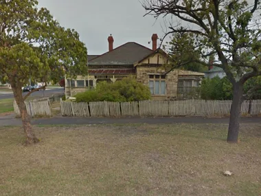 A weathered, single-story house with a picket fence and overgrown bushes in front, located on a street corner.