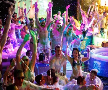 Colorful crowd in hot tubs enjoying a foam-filled cinema experience, under vibrant lighting at "Hot Tub Cinema."