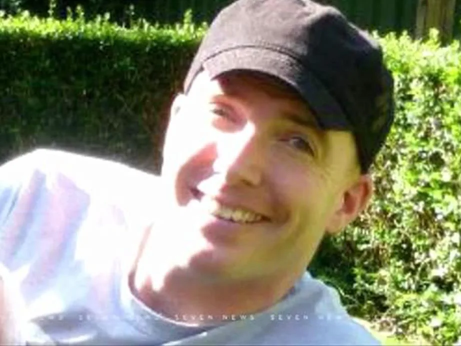 A man wearing a cap, smiling outdoors with greenery in the background.