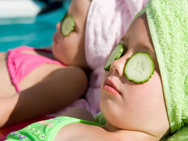 Two young girls sunbathing with cucumber slices over their eyes, wearing colorful swimsuits and towels on their heads.