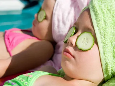 Two young girls sunbathing with cucumber slices over their eyes, wearing colorful swimsuits and towels on their heads.