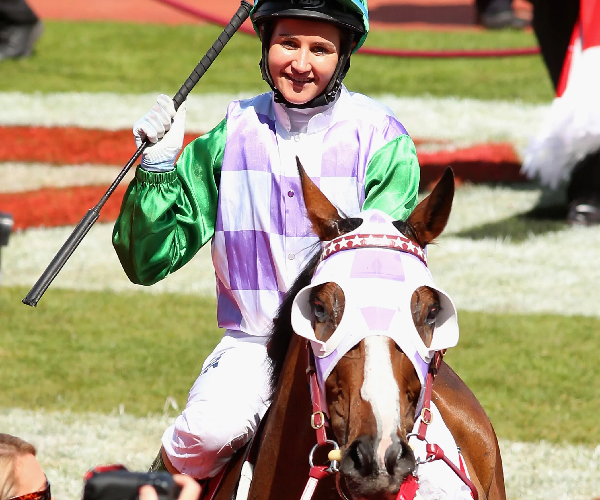 Jockey smiling, wearing green and purple silks, holding a whip, riding a horse with a white mask on a racetrack.
