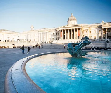 Trafalgar Square fountain with the National Gallery in the background under a clear blue sky.