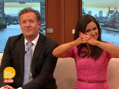 Two television hosts sitting on a sofa; one is wearing a suit and the other a pink dress with a playful expression.