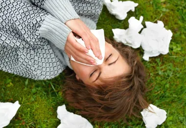 Person lying on grass blowing nose with tissues scattered around.