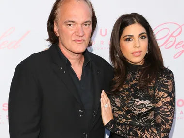 A man in a black suit poses with a woman in a lace dress at an event, standing against a white background with pink script.